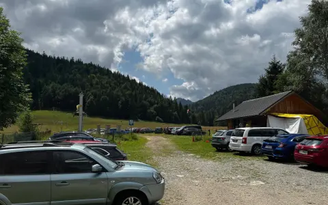 Parking z samochodami przy górskim schronisku, w tle las i chmury.