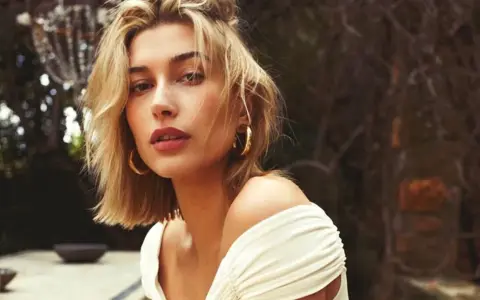Mujer con un moderno corte bob rubio, aretes dorados y un vestido blanco con hombros descubiertos, posando al aire libre.