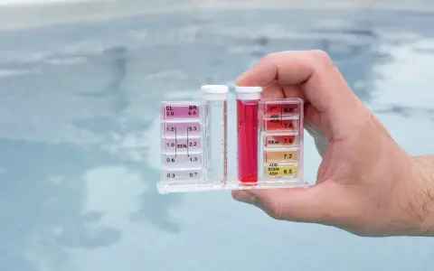 Hand holds pool water test kit. Red liquid in the pH tube indicates a high reading, suggesting acid is needed to lower pH.