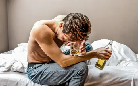 Mężczyzna siedzi na łóżku, trzymając szklankę z alkoholem i butelkę.