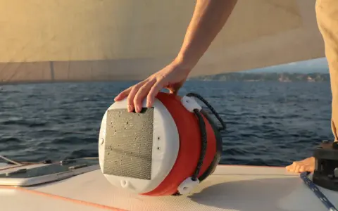 Main d'un marin tenant une bouée orange et blanche, un câble y est accroché. Le bateau navigue sur une mer bleue sous un ciel clair.