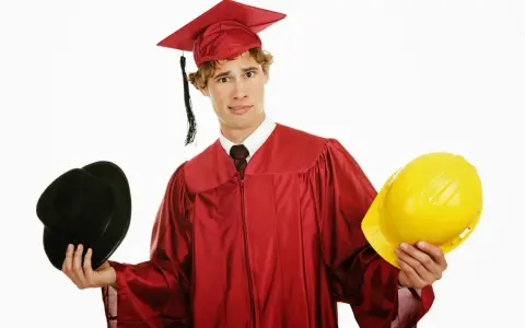 Studente universitario lavoratore con toga e cappello da laureato, tiene in mano un cappello nero e un casco giallo, indeciso sul futuro.