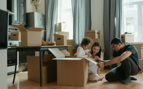 Rodzina pakuje kartony, przygotowując się do przeprowadzki. Dziecko rysuje na kartonie, a rodzice pomagają, planując jak się zameldować w nowym miejscu.