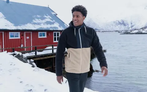 Mann in warmer Fleecejacke, bereit für das Waschen, steht vor einem roten Haus am Meer.