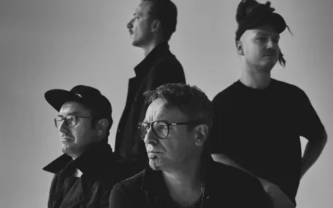 Czterech mężczyzn w czarnych ubraniach, spięty black metalowy zespół, z różnymi fryzurami i okularami, pozuje do zdjęcia.