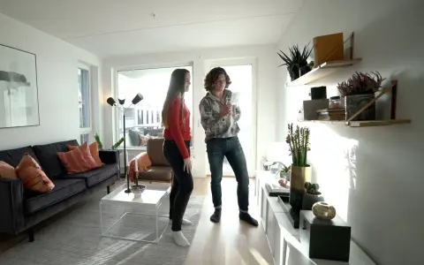 Två personer diskuterar något i ett modernt vardagsrum med trendig inredning, mörk soffa och hyllor med växter.