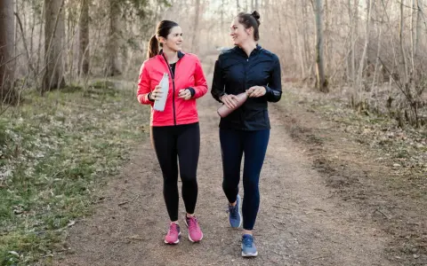 Two women in athletic wear walk on a forest path, holding water bottles.