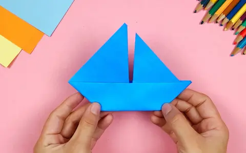 Hände falten ein blaues papierschiffchen. Bunte Stifte und Papier liegen bereit, um das papierschiffchen bauen zu beginnen.