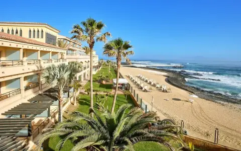 Fuerteventura kiedy wieje, palmy kołyszą się nad piaszczystą plażą i oceanem. Hotel z tarasami i widokiem na fale.