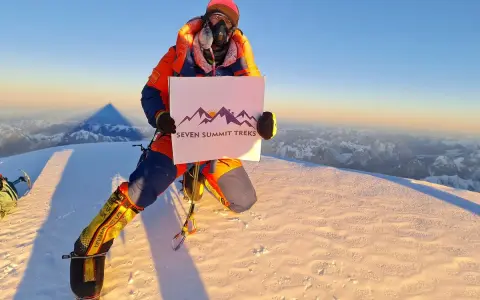 Na szczycie, gdzie jest K2, wspinacz w pomarańczowym kombinezonie trzyma tabliczkę "Seven Summit Treks".