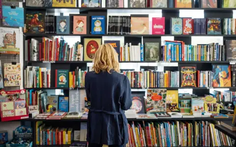 Donna bionda in libreria, circondata da scaffali pieni di libri colorati. Un'atmosfera di lavoro e scoperta tra le pagine.