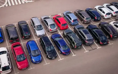 Rząd samochodów na parkingu, w tym czerwony, niebieski i srebrny. Jeden z nich czeka na odholowanie.