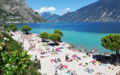 Spiaggia di Limone sul Garda affollata di bagnanti sotto le montagne. Acqua cristallina e sole splendente.