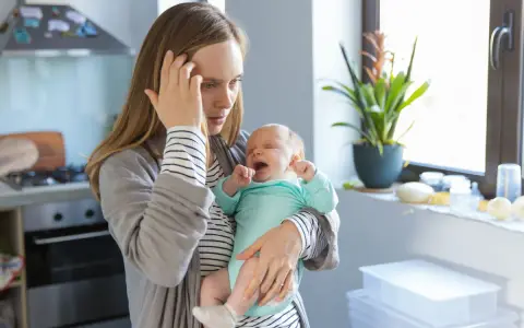 Müde Mutter hält weinendes Baby. Der **12 wochen schub** macht ihr zu schaffen. Sie hält sich die Stirn.
