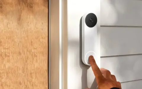 Un dedo presiona qué es el timbre inteligente, un dispositivo blanco con cámara negra, junto a una puerta de madera.