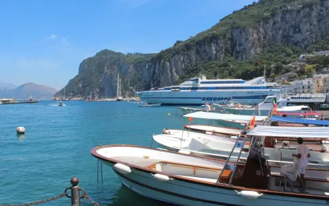 Barche in attesa al porto, con la maestosa isola di Capri sullo sfondo. Un traghetto Caremar attende passeggeri da Napoli.