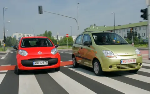 Czerwony i zielony samochód na pasach. Czy w terenie zabudowanym można wyprzedzać? Kierowcy zbliżają się do skrzyżowania.