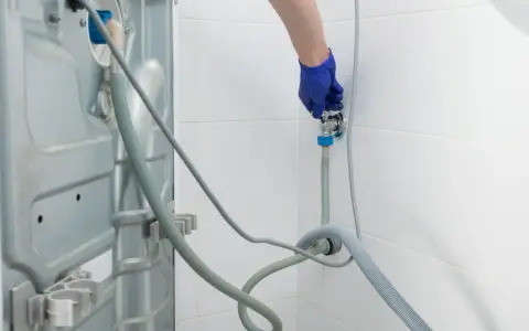 Blaue Handschuhe schließen den Wasserhahn für den Waschmaschinen Anschluss. Ein Schlauch führt zur Rückseite der Maschine.