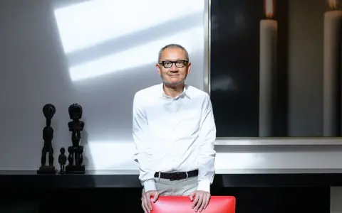 Pierre Chen, a man with glasses, stands in a modern room with striking shadows. He wears a white shirt and tan pants, leaning on a red chair.