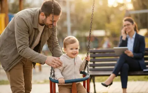 Ein Vater schaukelt sein Kind. Im Hintergrund telefoniert eine Frau. Die Frage, an welchen Kosten sich der Vater beteiligen muss, bleibt offen.