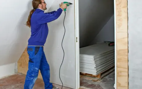 Man med skruvdragare monterar gipsskivor, kanske för att fixa en spackelremsa jula. Staplade skivor syns i bakgrunden.
