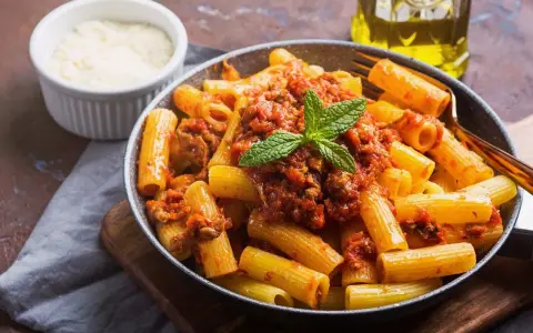 Un plato de macarrones con carne picada, cubiertos con salsa de tomate y decorados con menta. Al lado, un cuenco de queso rallado y una botella de aceite de oliva.