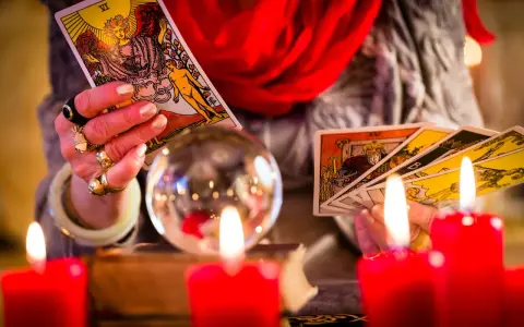 Dłoń z pierścieniami trzyma karty tarota, przygotowując tarot tygodniowy dla Bliźniąt. W tle kryształowa kula i czerwone świece.