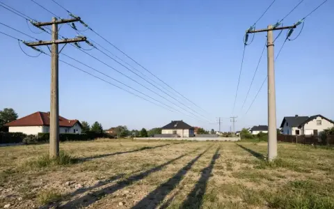 Dwa słupy energetyczne z przewodami linii wysokiego napięcia 110 kV, nad pustą działką z domami w tle. Zachowanie bezpieczna odległość od linii wysokiego napięcia 110 kv jest kluczowe.