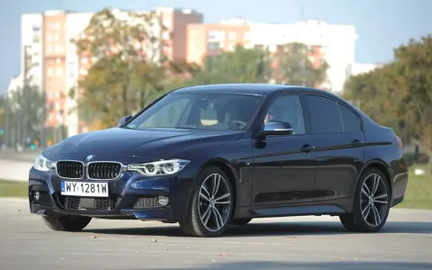 Ciemnoniebieskie BMW 318d z polskimi tablicami rejestracyjnymi stoi na tle budynków i drzew.