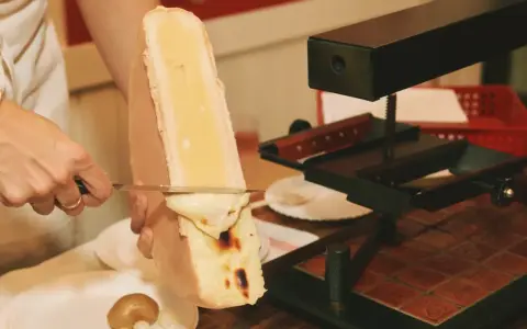 En person skrapar smält ost från en stor ostbit med en kniv. Perfekt för en mysig kväll med vin till raclette.