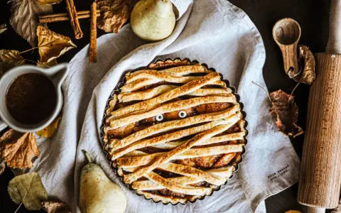 Tarte d'Halloween en forme de momie, avec des yeux globuleux et une garniture aux fruits. Autour, des feuilles d'automne, des poires et des épices.
