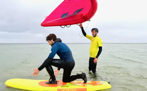 Zwei Männer üben Wing Surfen im flachen Wasser. Einer kniet auf einem gelben Board, der andere hält einen roten Wing.