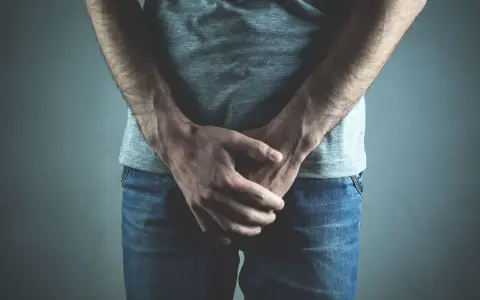 Mężczyzna w dżinsach i szarej koszulce, ręce skrzyżowane na kroczu. Może to symbolizować dyskomfort lub problemy zdrowotne, takie jak wodniak jądra.