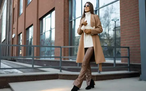 Elegancka kobieta w płaszczu w camelowy kolorze i brązowych spodniach ze skóry ekologicznej pozuje na tle nowoczesnego budynku.