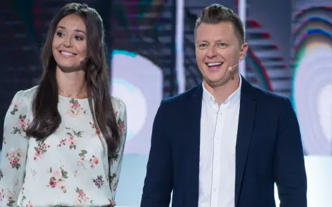 Uśmiechnięta para na scenie, gdzie nagrania programu telewizyjnego. Ona w kwiecistej sukience, on w marynarce.