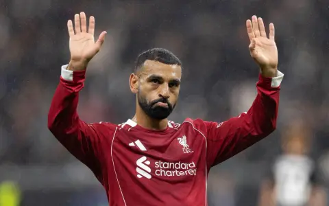 Mohamed Salah w czerwonej koszulce Liverpoolu, z uniesionymi dłońmi, pozdrawia kibiców. Widać logo klubu i sponsora.