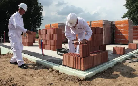 Dwóch budowlańców w białych kombinezonach układa cegły na fundamencie.