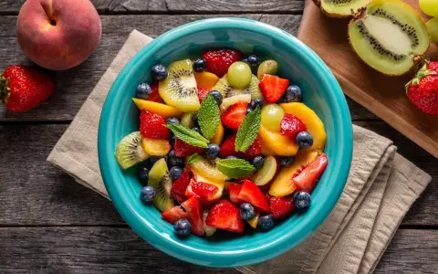 Un bol turquoise débordant de salade de fruits été : fraises, myrtilles, kiwis, pêches et raisins, garnie de menthe fraîche.