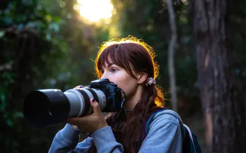 Młoda kobieta z aparatem w lesie, ćwiczy na zajęciach szkoły fotografii. Słońce przebija się przez drzewa.