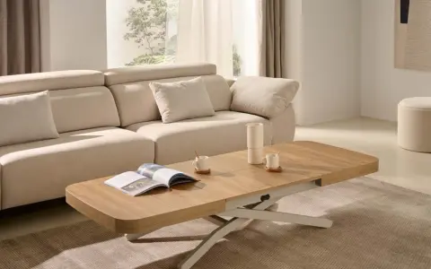 Mesa centro elevable 120x70 de madera clara, con un libro abierto y taza de café, junto a un sofá beige en un salón luminoso.