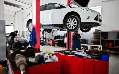 Warsztat samochodowy: biały samochód na podnośniku, mechanik w niebieskim kombinezonie.