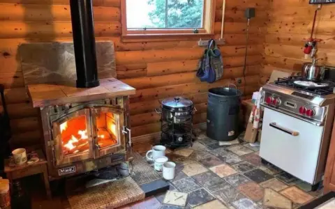 Poêle à bois allumé dans un chalet en bois rond, respectant la norme d'installation. Cuisine fonctionnelle avec cuisinière et ustensiles.