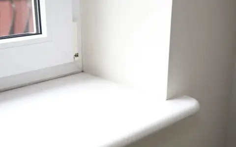 Biały parapet okienny, gotowy do wymiany. Widoczny fragment okna i ściany.