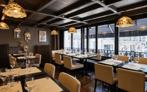 Salle à manger élégante avec des tables dressées, des luminaires Tiffany et une affiche "Les Artistes Limoges Menu" au mur.