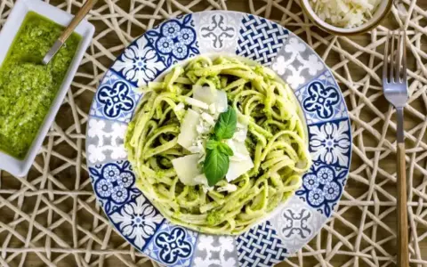 Pasta al pesto servida en plato decorado, con salsa de pesto y queso rallado al lado.