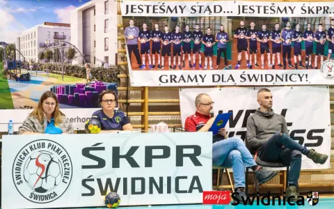 Na banerze widnieje drużyna SKPR Świdnica. Dwie kobiety siedzą za stołem z piłką ręczną.