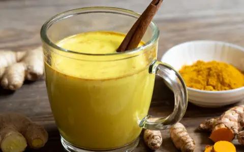 Taza de leche dorada con canela, rodeada de raíz de cúrcuma fresca y en polvo.