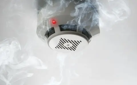Biały czujnik dymu na suficie, otoczony kłębami dymu. Czerwona dioda sygnalizuje alarm. Tak wygląda czujnik dymu w akcji.