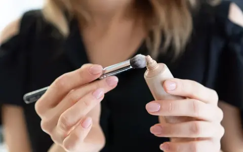 Dłonie z perfekcyjnym manicure nakładają pędzelkiem dr irena eris podkład rozświetlający na pociągłą tubkę.