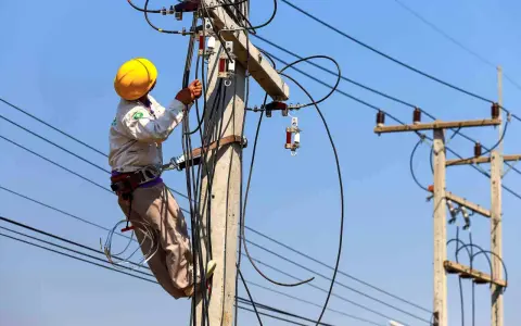 Elektryk w żółtym kasku pracuje na słupie energetycznym, podłączając przewody. Zastanawiasz się, ile kosztuje przyłącze elektryczne?